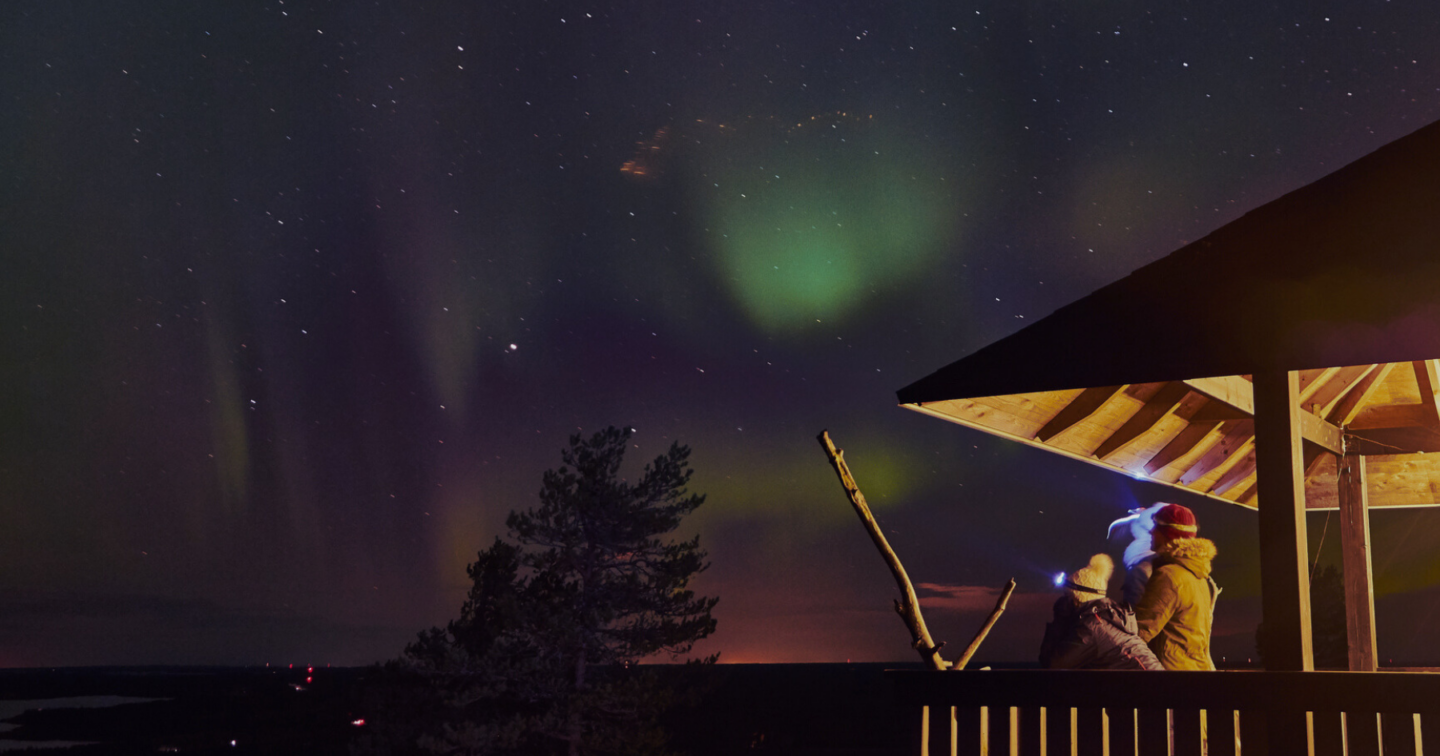 Two people look at a star filled sky from a warm lit deck at night.